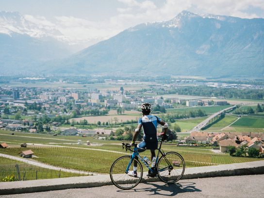 Een man kijkt zittend op de stang van zijn fiets over een bebouwd landschap met op de achtergrond bergen.