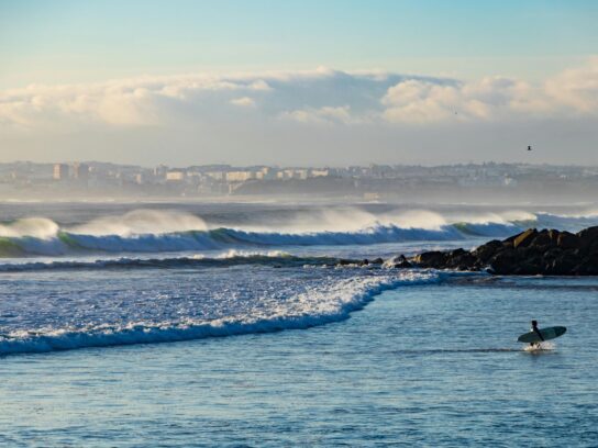 Persoon met een surfboard in de zee met op de achtergrond de kustlijn van Costa da Caparica.